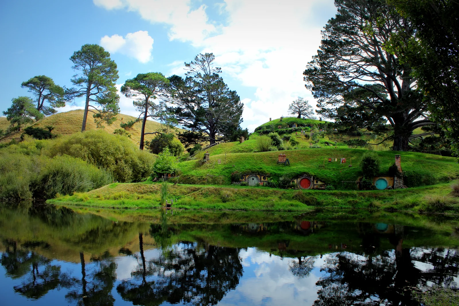 Casas de portas redondas no Hobbiton Movie Set, entre colinas verdes e árvores, refletidas em um lago.