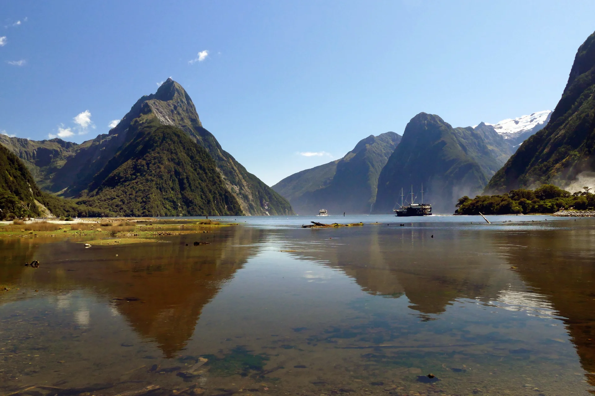 Montanhas de Milford Sound refletidas nas águas do fiorde, com barcos ao fundo.